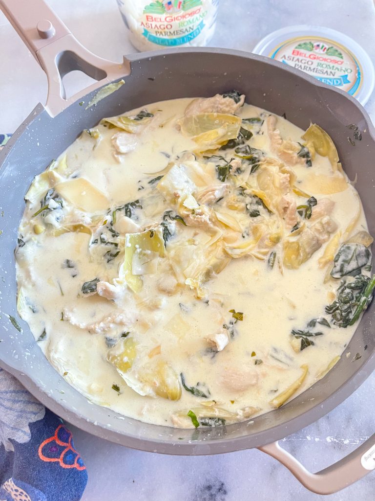 This is an image of a light brown pan with creamy lemon spinach and chicken with a tub of Parmesan cheese in the background.