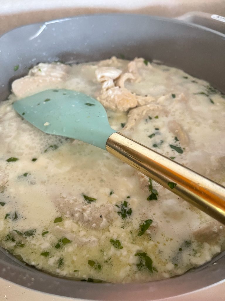 Creamy lemon artichoke chicken cooking in brown pan with blue and gold rubber spatula.