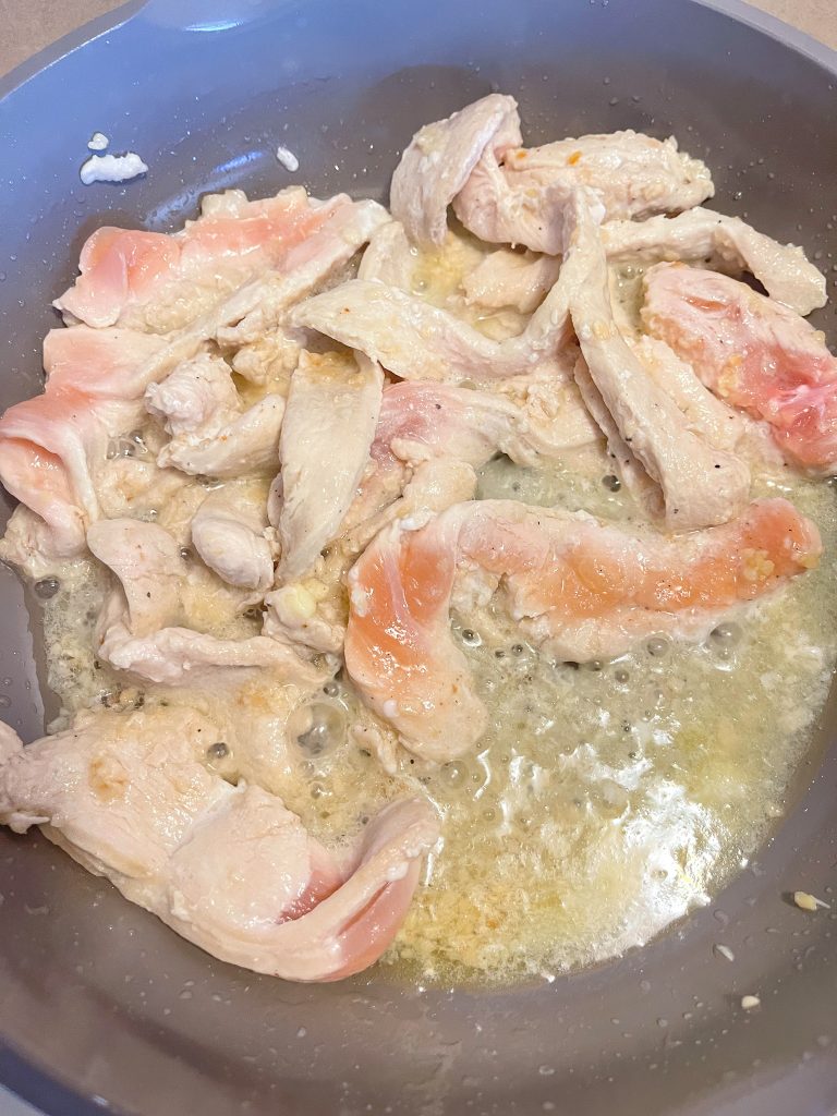 Sliced chicken breasts cooking in avocado oil with minced garlic in brown pan.