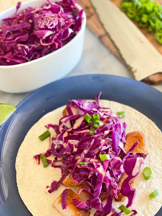 Image depicts a blue plate with a tortilla, blackened chicken spicy red cabbage slaw and green onion. A lime on the side of the plate and a white bowl with cabbage slaw on a gray table with a cutting knife in the background.