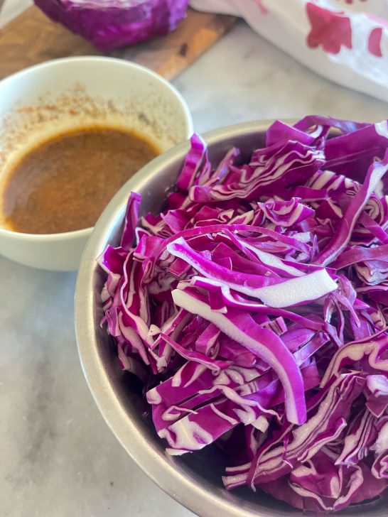 Image depicts a bowl of shredded red cabbage and a white bowl of brown liquid sauce on a grey table.
