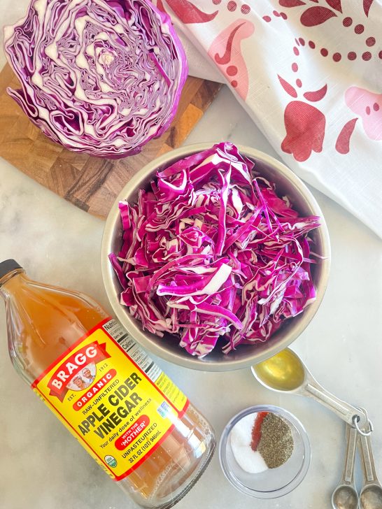 Image depicts half a red cabbage on a wooden cutting board, a white and red tea towel, a silver bowl of shredded red cabbage, a jar of apple cider vinegar, a tablespoon of olive oil, and a small container with hot sauce, salt, sugar and black pepper in it on a gray background.