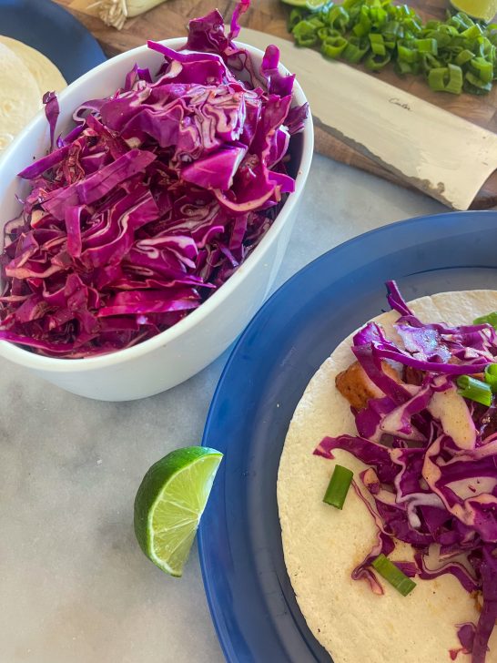 Image depicts a blue plate with a tortilla, blackened chicken spicy red cabbage slaw and green onion. A lime on the side of the plate and a white bowl with cabbage slaw on a gray table.