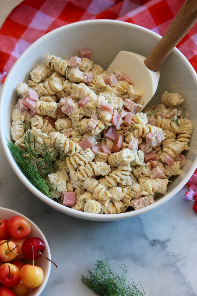 Image depicts white serving bowl filled with macaroni salad with dill and ham and a small bowl of cherries in the foreground on a gray counter top