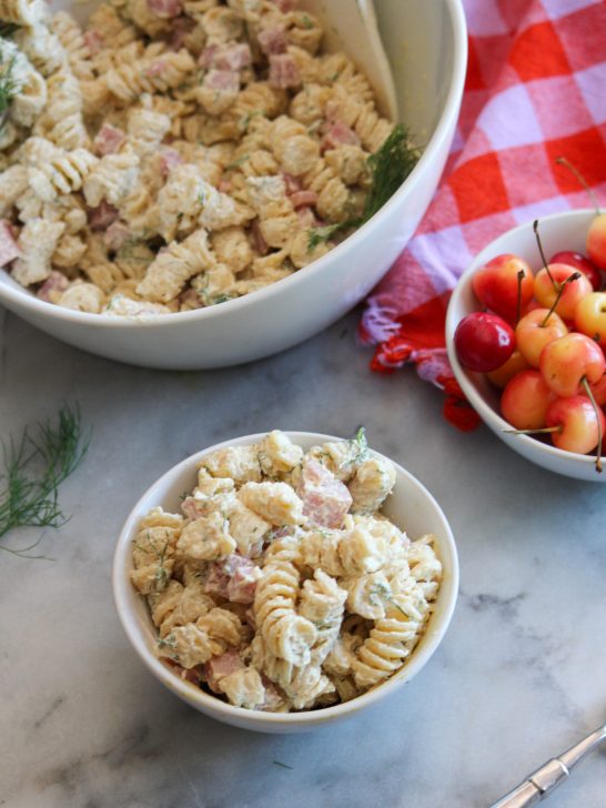Image depicts white bowl of Danish Macaroni Salad with a smaller dish of it in front. A bowl of cherries and a purple and red dish towel on a gray background.