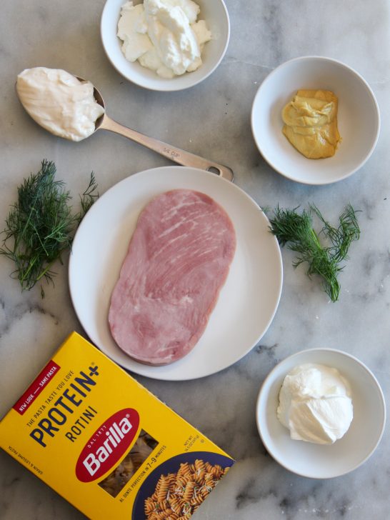 Image depicts box of protein rotini, sprigs of fresh dill, a slice of ham steak, some sour cream and yogurt in white dishes and Dijon mustard in a white dish on a gray counter top.